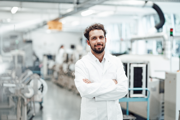 Selbstbewusster männlicher Wissenschaftler in weißem Laborkittel, der mit verschränkten Armen in einem hellen Labor steht