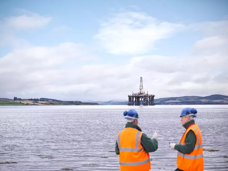 Two male oil worker on shore with oil rig in background