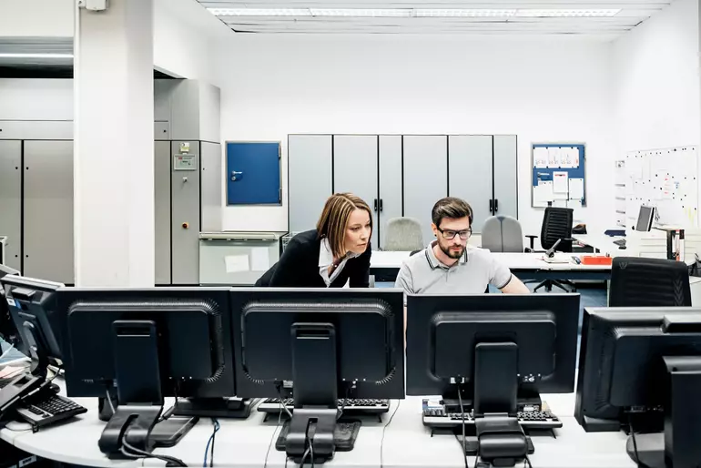 Eine Betriebsleiterin einer Druckerei, die ihren Kollegen bei der Arbeit an mehreren Computern im Kontrollraum unterstützt
