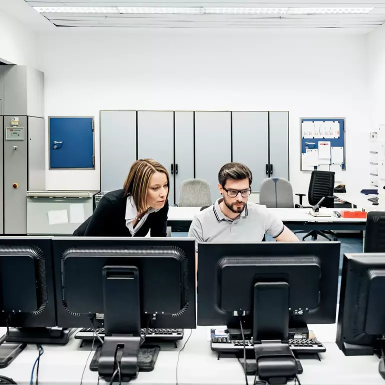 Eine Betriebsleiterin einer Druckerei, die ihren Kollegen bei der Arbeit an mehreren Computern im Kontrollraum unterstützt