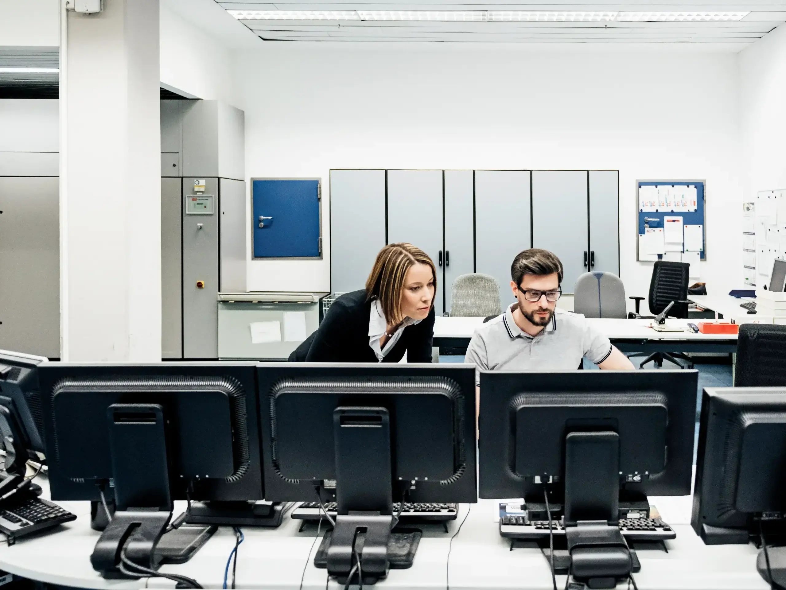 A printing factory operations manager assisting her colleague working at multiple computers in the control room