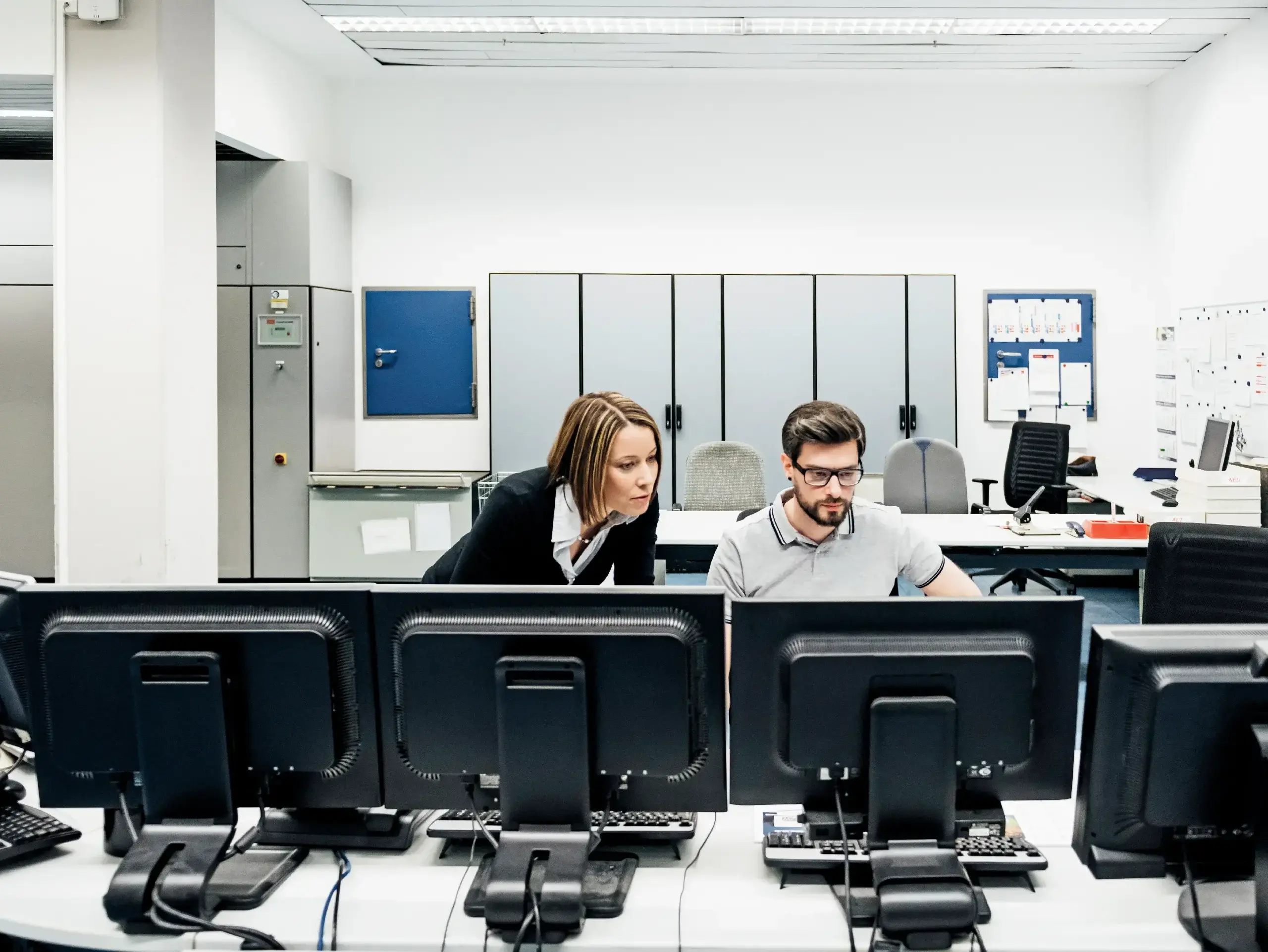 Eine Betriebsleiterin einer Druckerei, die ihren Kollegen bei der Arbeit an mehreren Computern im Kontrollraum unterstützt