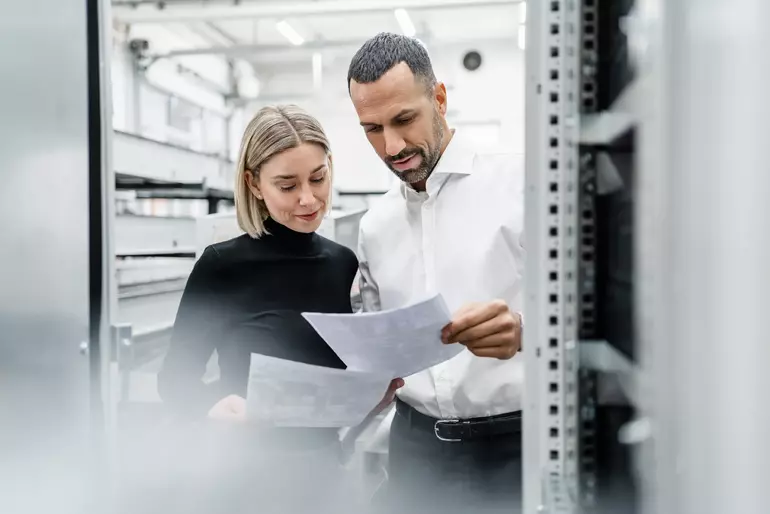 Businessman and woman with papers at a machine in factory hall